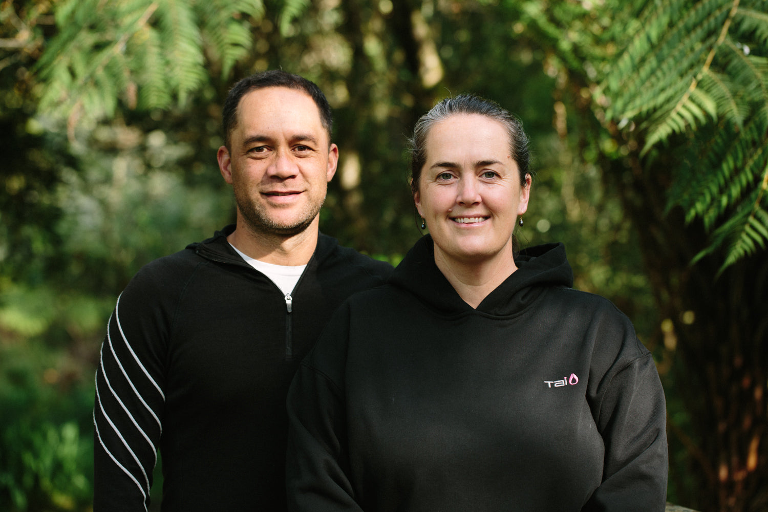 Two people wearing black hoodies standing in front of green foliage